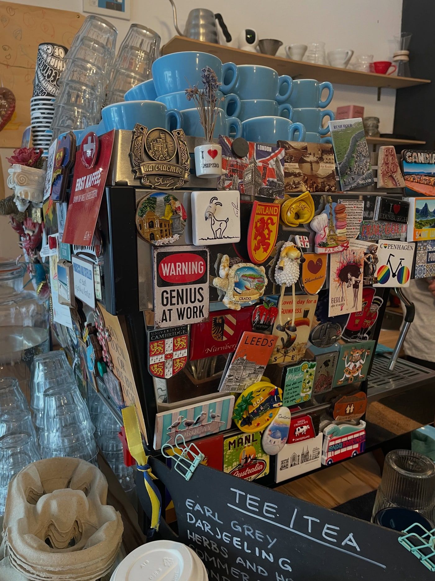 A shelf of blue cups above a wall of fridge magnets and a 'Tee / Tea' chalkboard