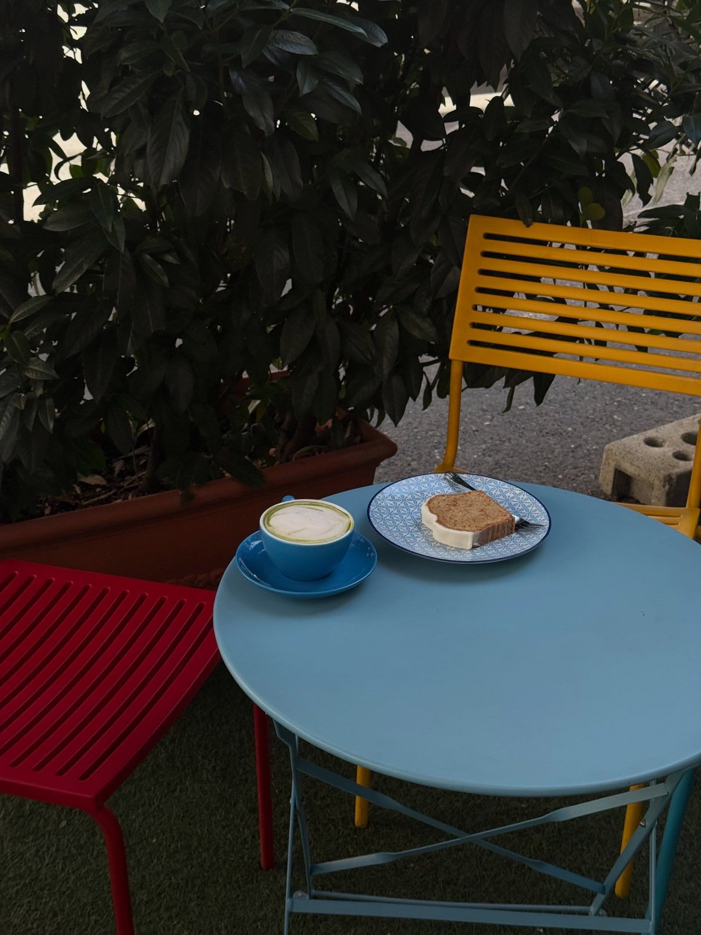 A cappuccino and cake on a blue table among yellow and red chairs outside